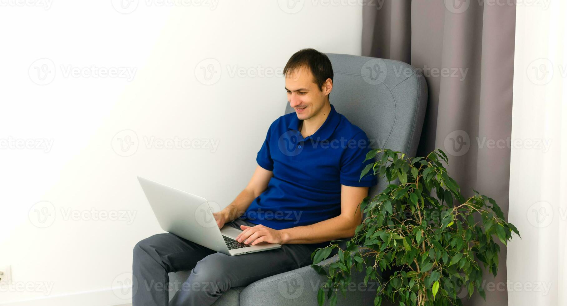 Happy young man in t-shirt sitting on sofa at home, working on laptop computer, smiling. photo