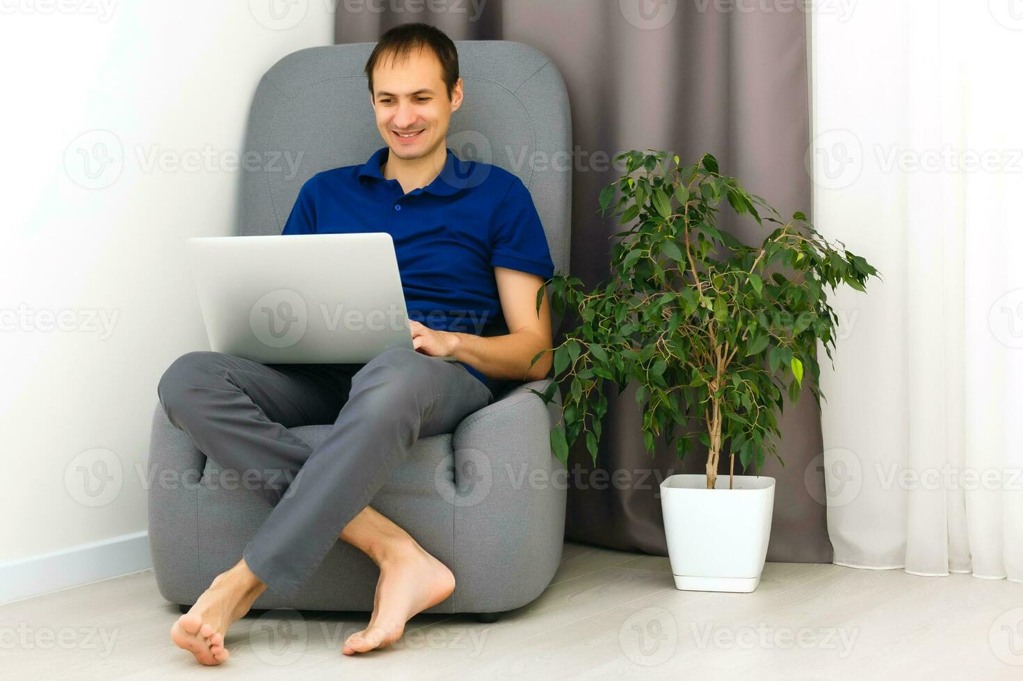 Happy young man in t-shirt sitting on sofa at home, working on laptop computer, smiling. photo