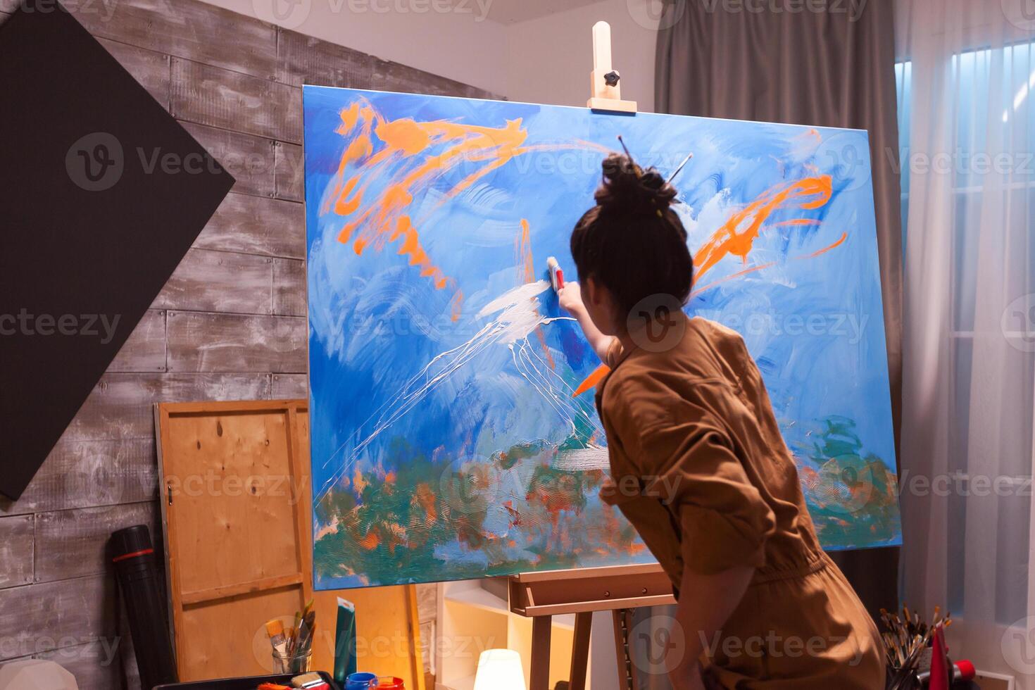 Skillful woman painter in art workshop painting on large canvas with orange color. Modern artwork paint on canvas, creative, contemporary and successful fine art artist drawing masterpiece photo