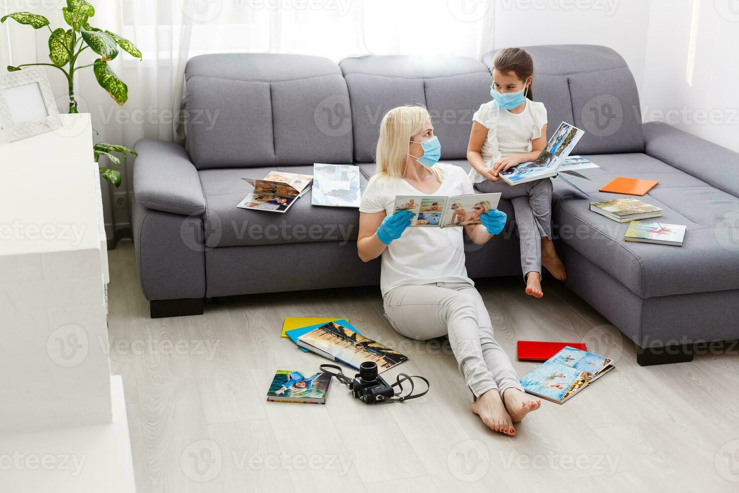 Quarantine time mother and little daughter in protective masks looking family photos album at living room together. photobook