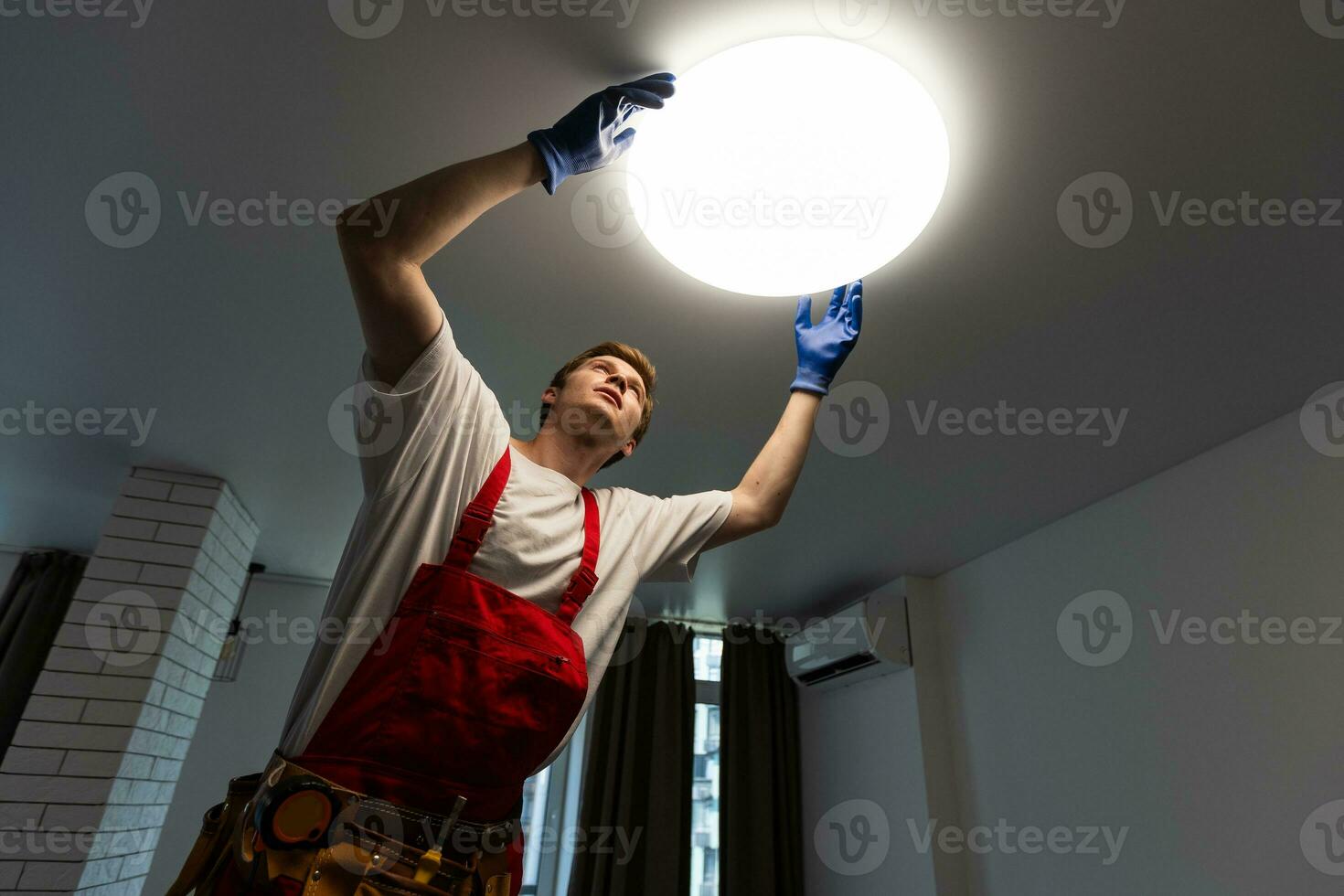 Electrician at work. Service for the repair of electrical wiring and replacement of ceiling lamps. A builder is installing a loftstyle wooden ceiling. Rentagent helps with the housework photo