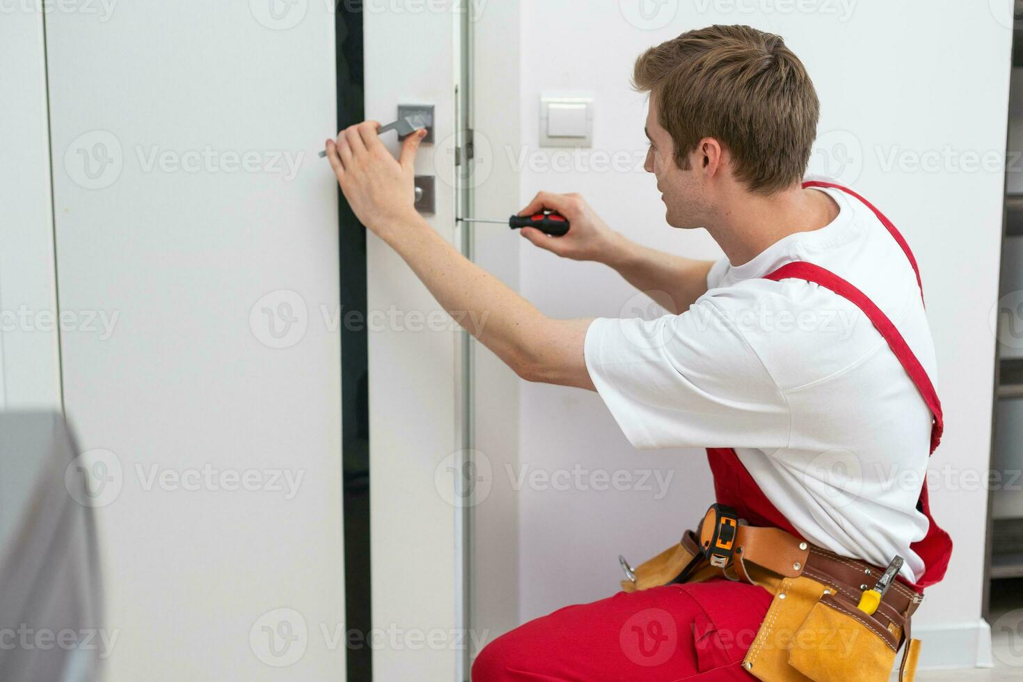 Installation of a lock on the front wooden entrance door. Portrait of young locksmith workman in blue uniform installing door knob. Professional repair service. Maintenance Concept photo