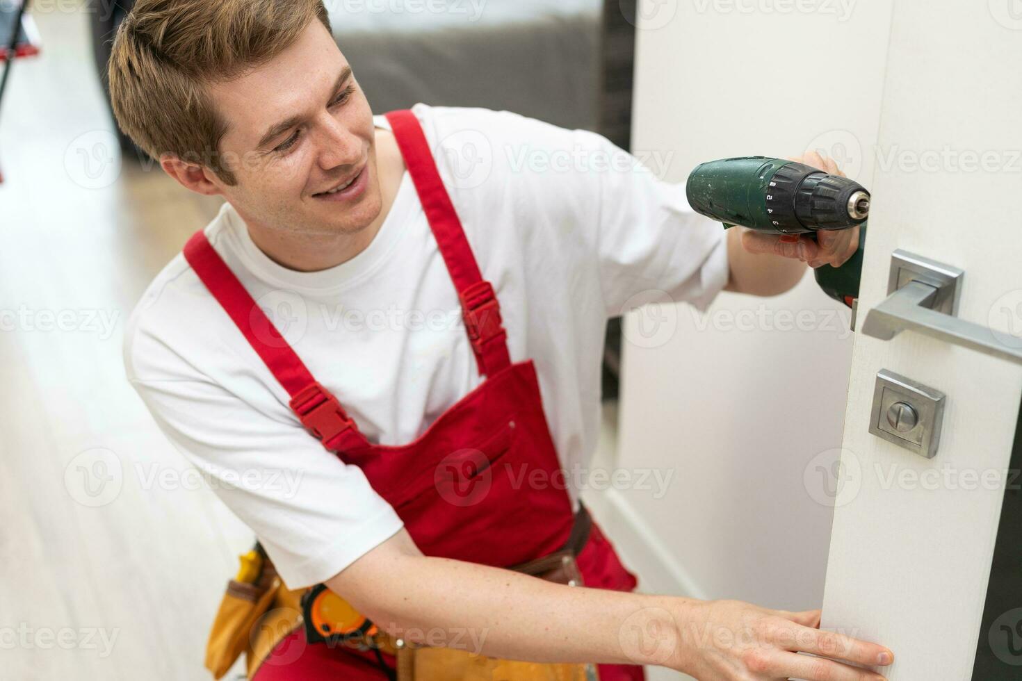 Installation of a lock on the front wooden entrance door. Portrait of young locksmith workman in blue uniform installing door knob. Professional repair service. Maintenance Concept photo