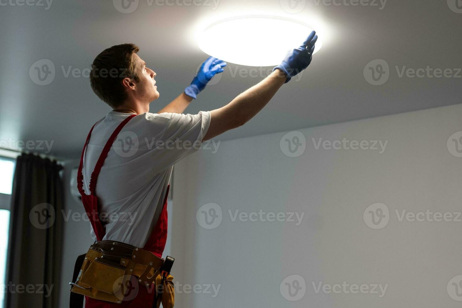 Electrician in uniform installing ceiling lamp indoors. Space for text photo