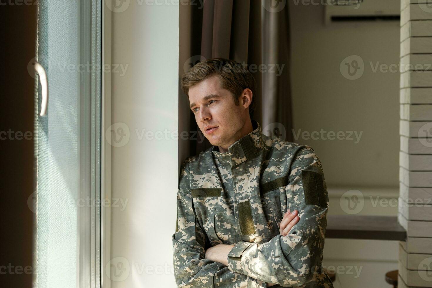 Depressed and sad soldier in green uniform with trauma after war standing near the window photo