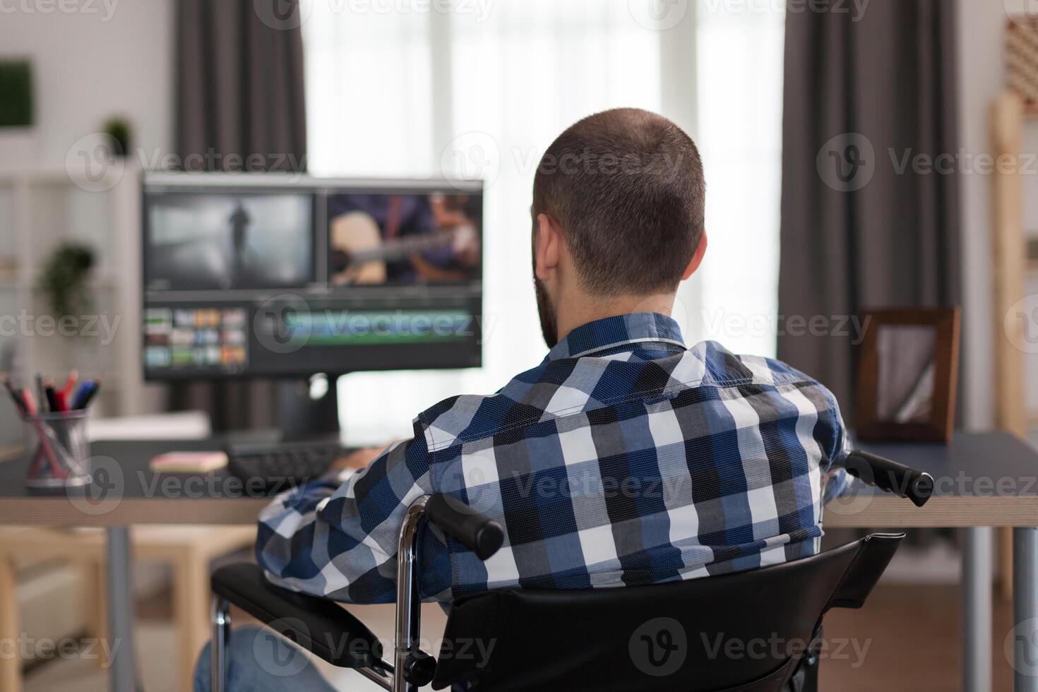 Disabled filmmmaker in wheelchair working on post production of a new video. Invalid videographer doing his business online, using high technology, sitting in his apartment, working remotely in special conditions. photo