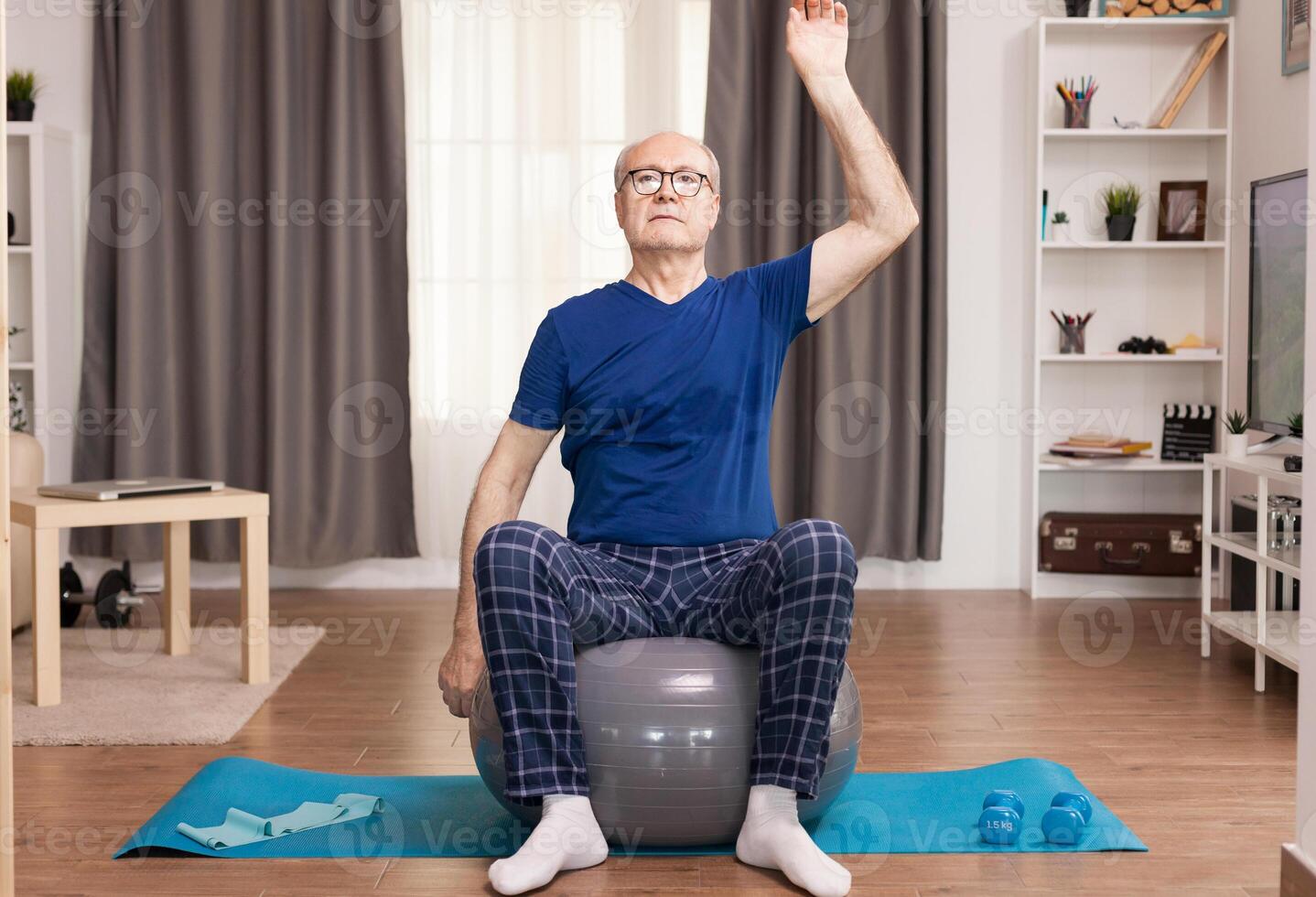 Elderly man training at home, doing arm exercises. Old person pensioner online internet exercise training at home sport activity with dumbbell, resistance band, swiss ball at elderly retirement age. photo