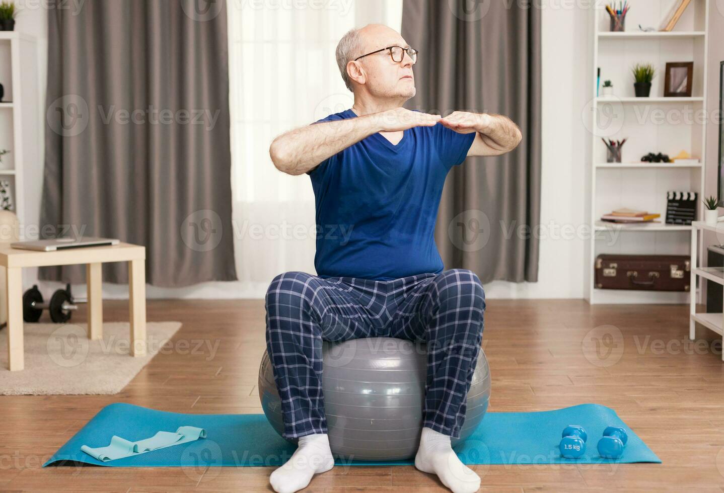 Active old man doing sport in his cozy apartment using swiss ball and yoga mat. Old person pensioner online internet exercise training at home sport activity with dumbbell, resistance band, swiss ball at elderly retirement age. photo