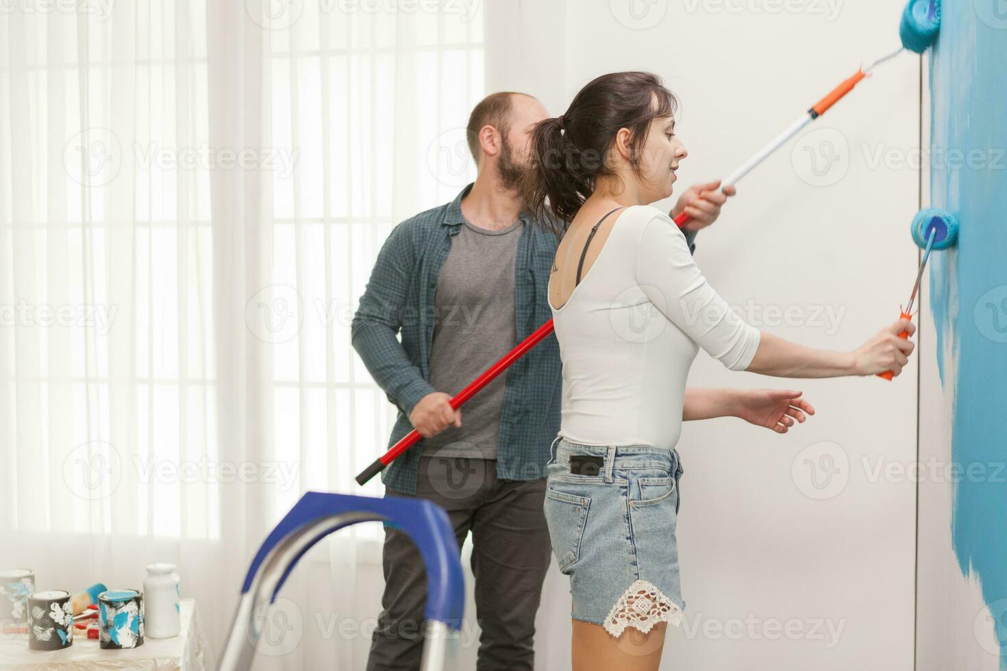 Happy couple painting together the apartment wall with rollers. Home during renovation, decoration and painting. Interior apartment improvement maintenance. Roller, ladder for house repair photo