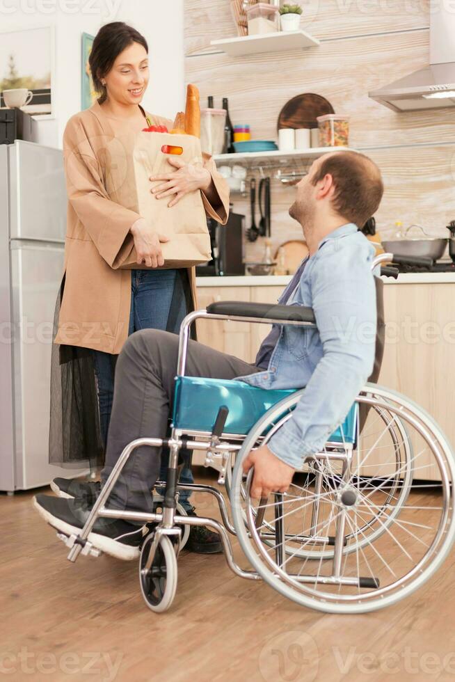 Wife holding paper bag with organic products in kitchen talking with disabled husband in wheelchair. Disabled paralyzed handicapped man with walking disability integrating after an accident. photo