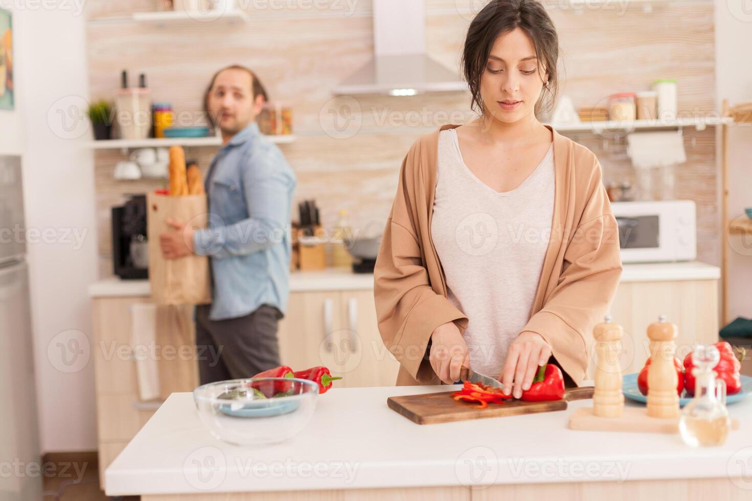 Wife cutting salad ingredient while husband is holding grocery bag with fresh vegetables from market. Funny happy in love couple at home spending time together, cooking healthy and smiling photo
