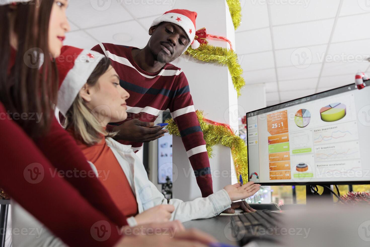 Meticulous business team checking financial statistics on computer screen during Christmas season. Teamworking employees analyzing data graphs and charts in festive decorated office photo