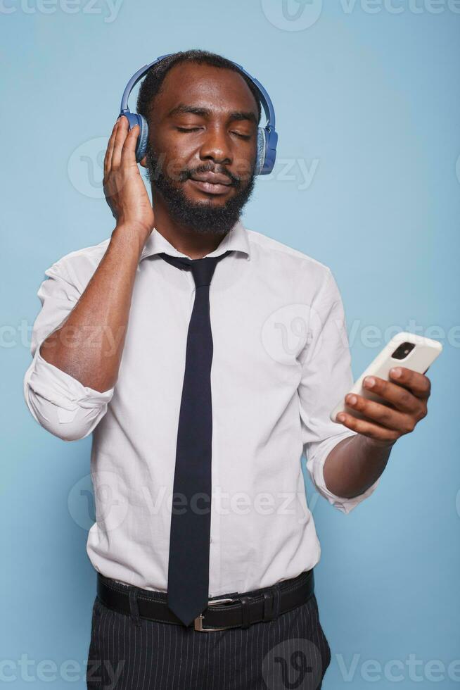 retrato de negro hombre con ojos cerrado disfrutando audio contenido transmisión desde teléfono inteligente a inalámbrico auriculares. joven oficina trabajador escuchando a calmante podcast en inalámbrico auriculares. foto