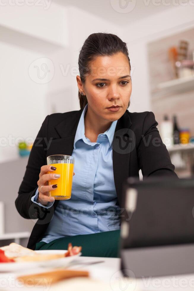 Young businesswoman holding a glass of juice and using the tablet in the morning. Freelancer reading the last news online before going to work, using modern technology in the kitchen while eating a healthy meal photo