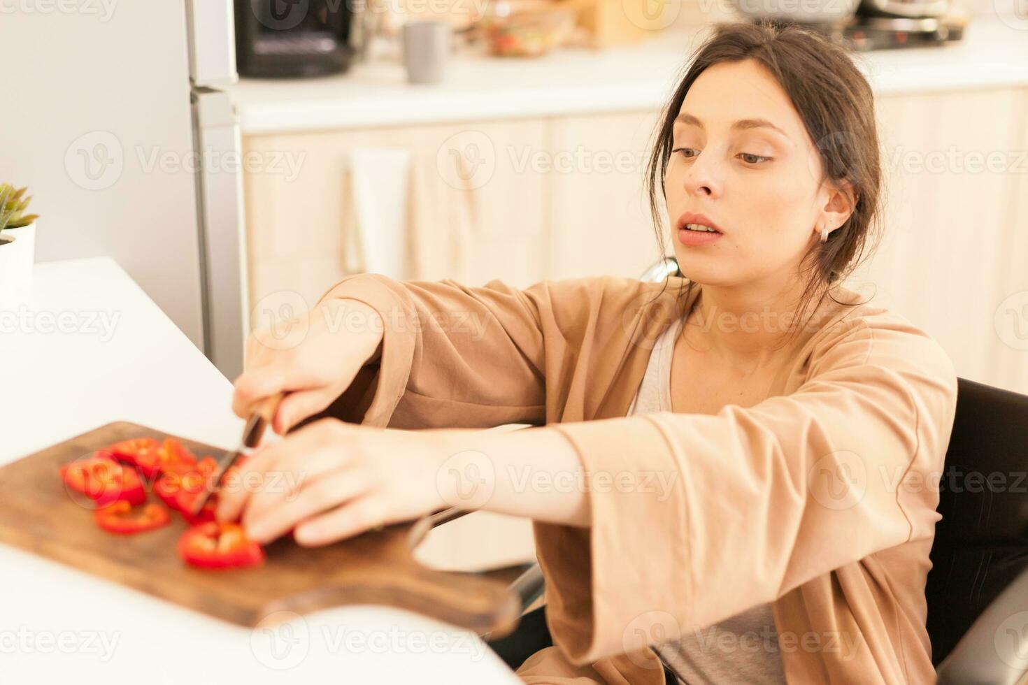 Woman with walking handicap cutting red paper on wooden board in kitchen using sharp knife. Disabled paralyzed handicapped woman with walking disability integrating after an accident. photo