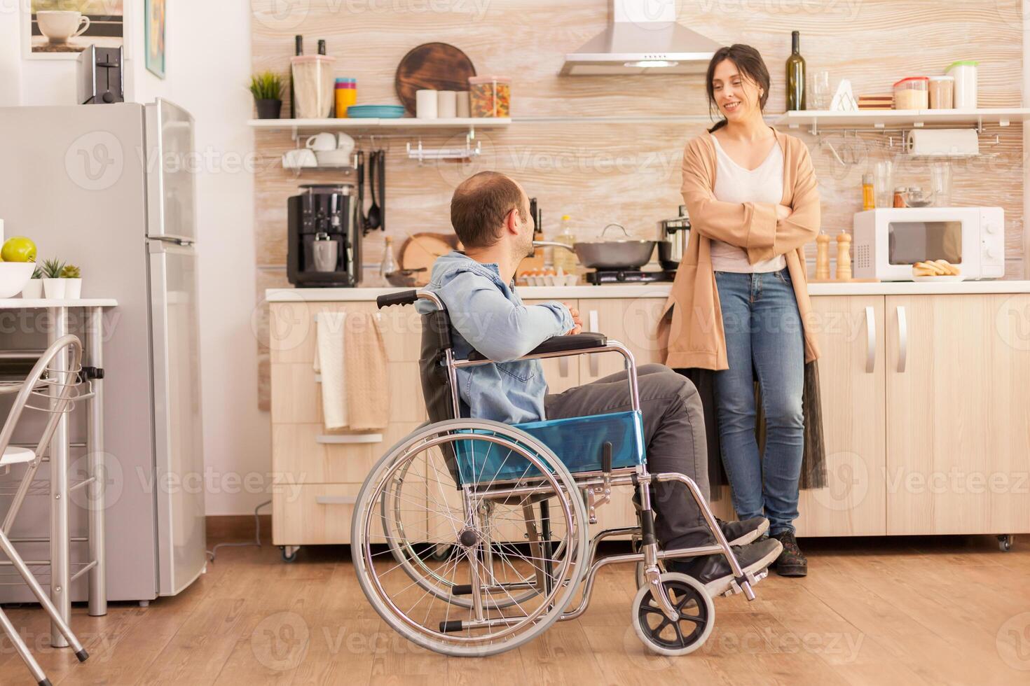 Happy wife in kitchen talking with disabled husband in wheelchair. Disabled paralyzed handicapped man with walking disability integrating after an accident. photo