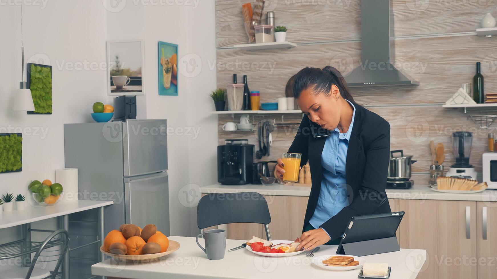 Businesswoman on phone while eating breakfast. Concentrated business woman in the morning multitasking in the kitchen before going to the office, stressful way of life, career and goals to meet photo