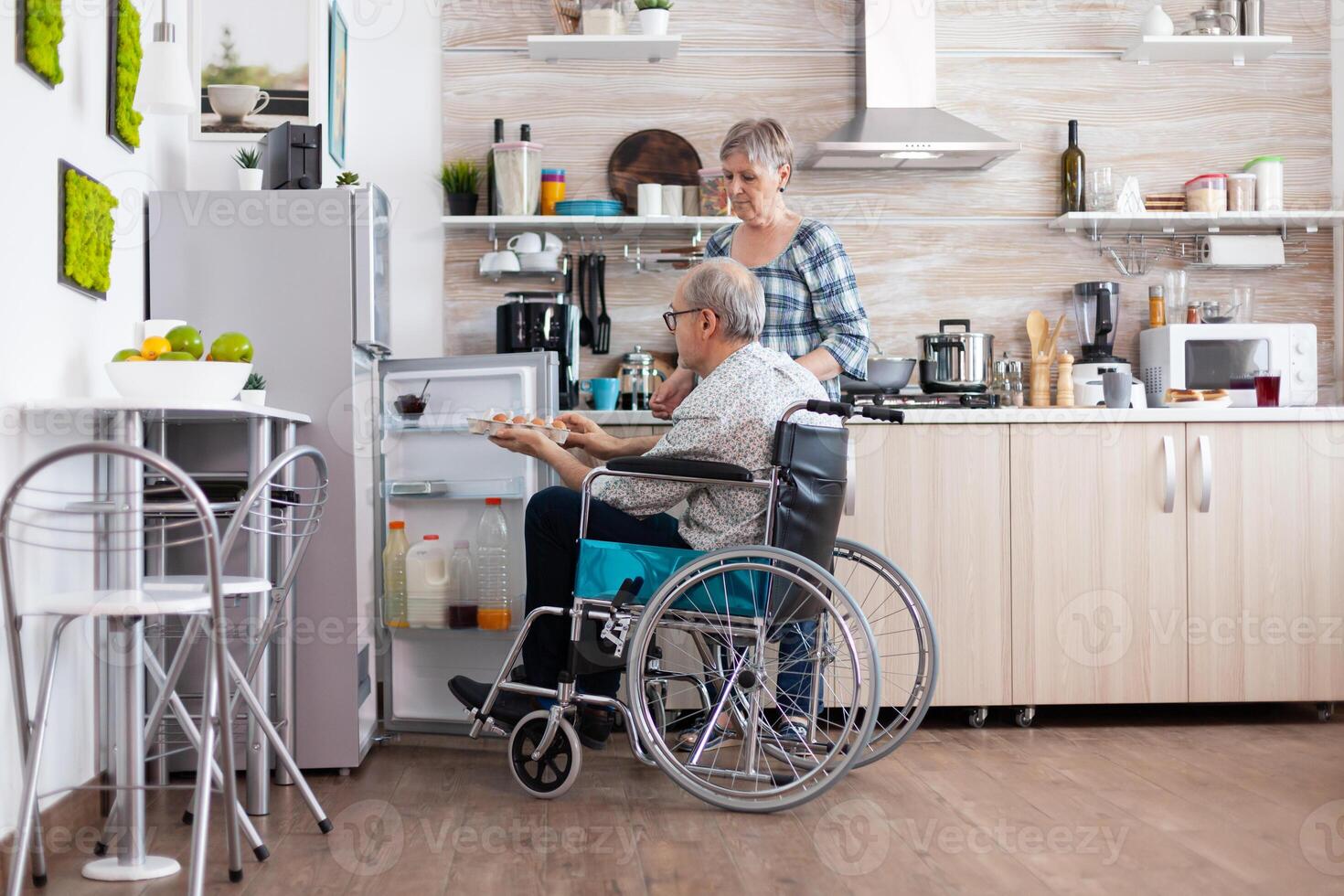 Disabled senior male in wheelchair taking eggs carton from refrigerator helping his wife in kitchen. Senior woman preparing breakfast for handicapped husband, living with man with walking disabilities photo