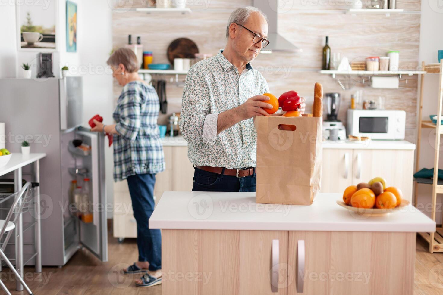 Cheerful happy family healthy lifestyle putting fresh fruits and groceries in refrigerator. Elderly husband and wife taking out vegetables from grocery paper bag after arriving from supermarket. photo