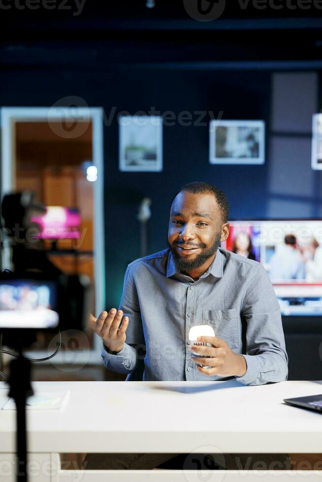 Influencer talking about house improvement video topic, showing newly released smart light bulb to viewers. Smiling african american tech fan presenting WIFI home automation photo