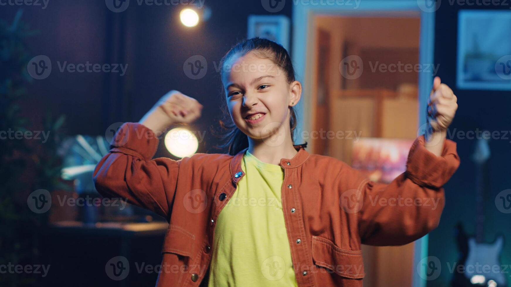 Close up shot of small girl doing captivating dance moves for social media platform, doing viral challenge. Cute little kid recording video for online fans in dimly lit home studio photo