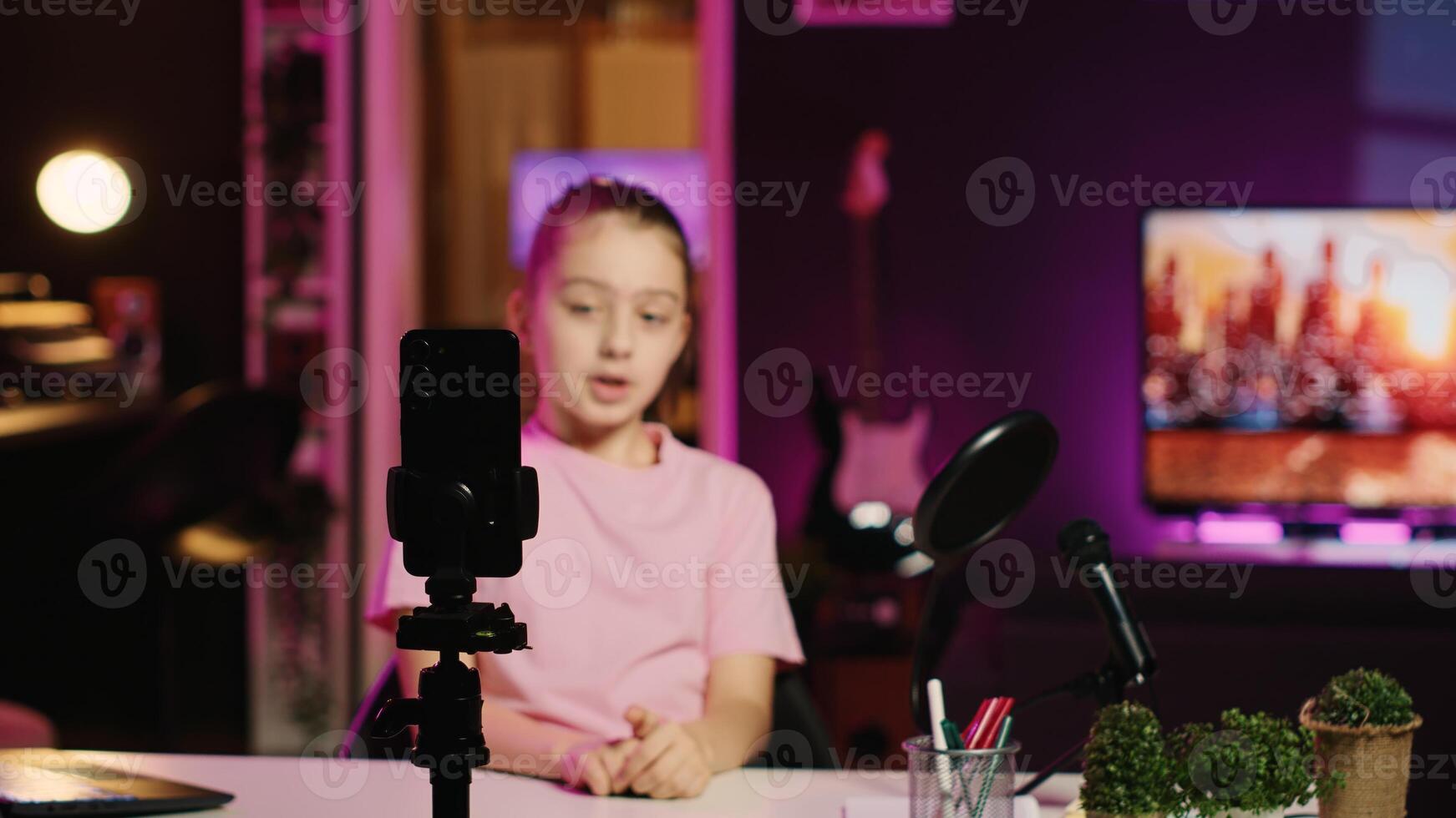 Focus shot on phone attached to tripod used for recording family vlog with happy child in blurry background talking about school activities with her children audience, filming content for social media photo