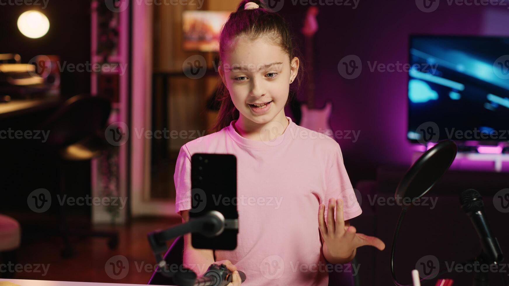 Kid films with smartphone attached to selfie stick in pink neon lit living room used as professional studio. Young media star captures footage with cellphone camera, discussing about fun day at school photo