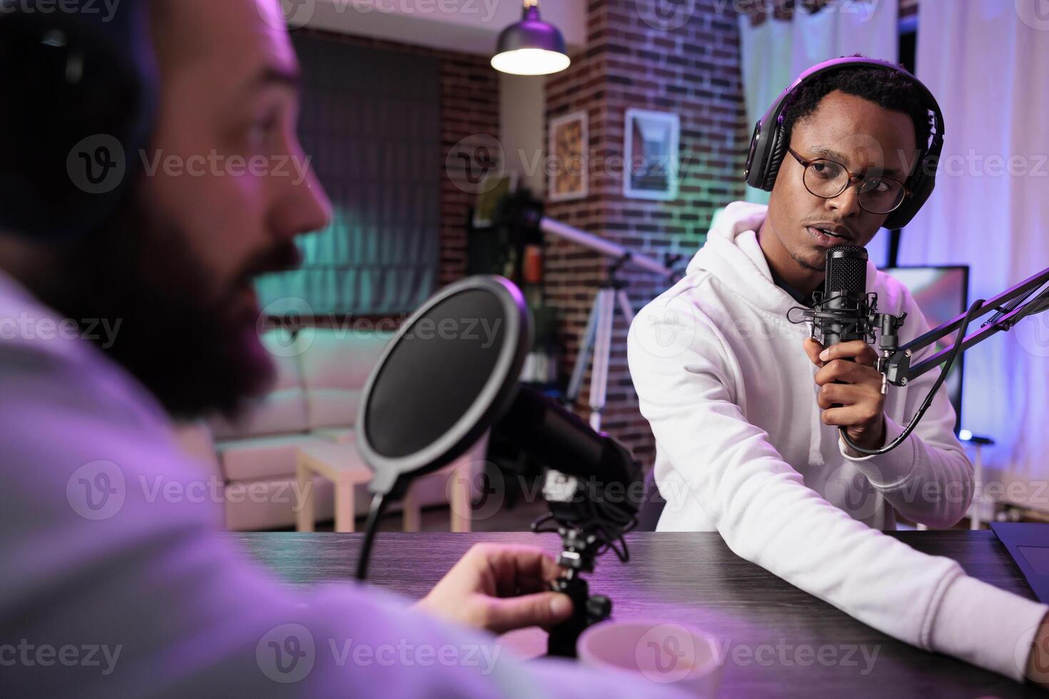 Wise guest on online podcast show telling inspiring story to host, entertaining viewers watching during live broadcast. Presenter using audio recording equipment in rgb lights decorated living room photo