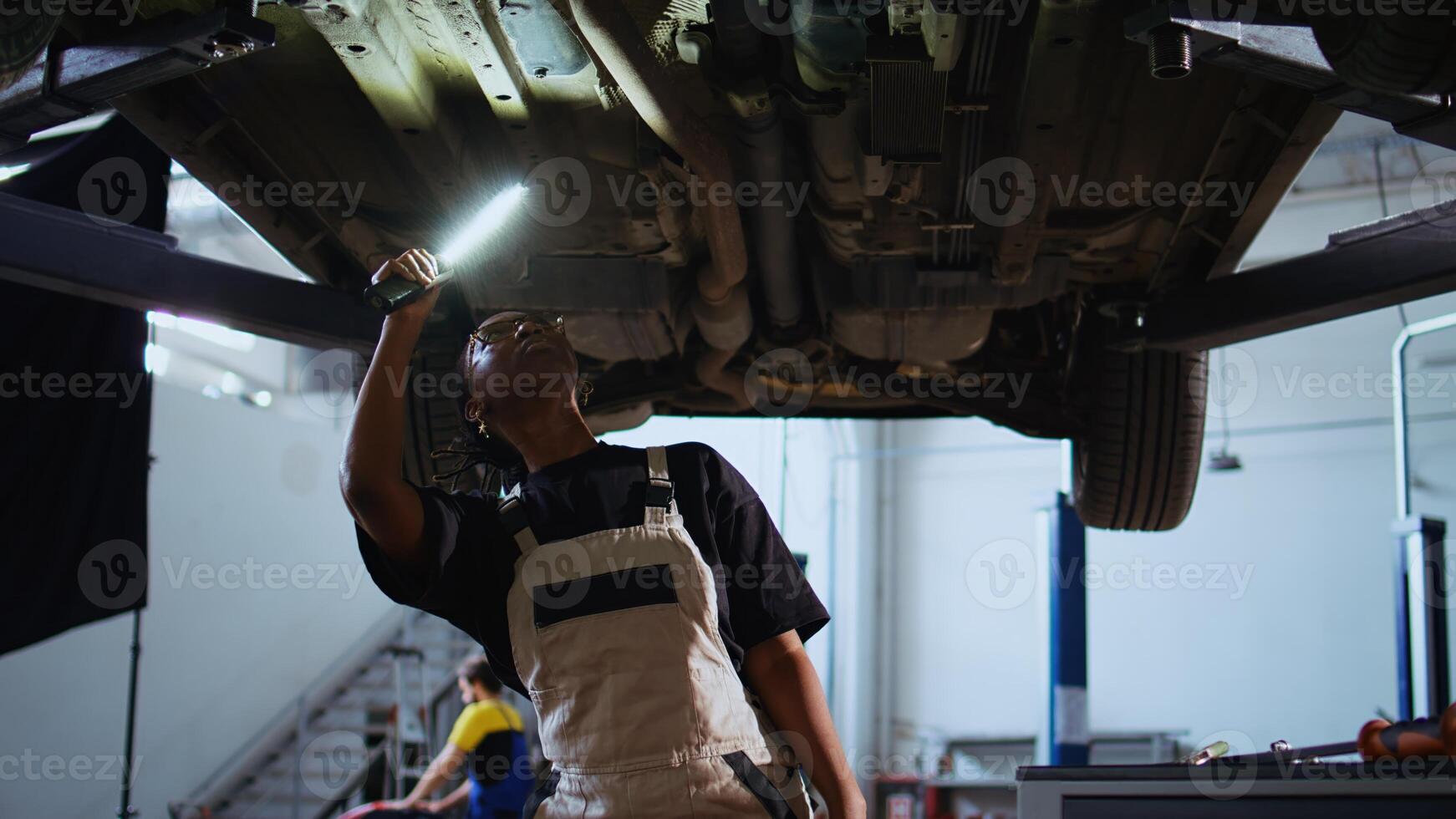 Trained repairman working on suspended car in garage, checking components during routine maintenance. African american repair shop worker walking underneath vehicle, examining it using work light photo