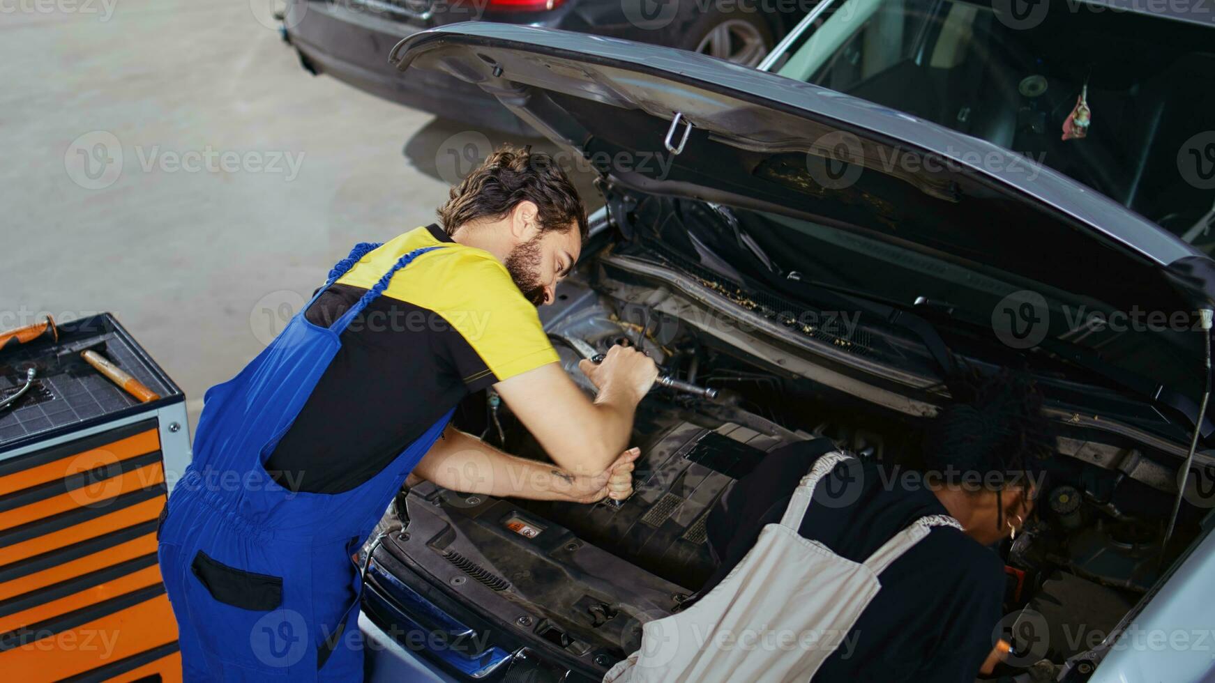 Qualified mechanic in garage using laptop to follow checklist while doing maintenance on car, talking with client. Employee in repair shop does checkup on automobile next to customer photo