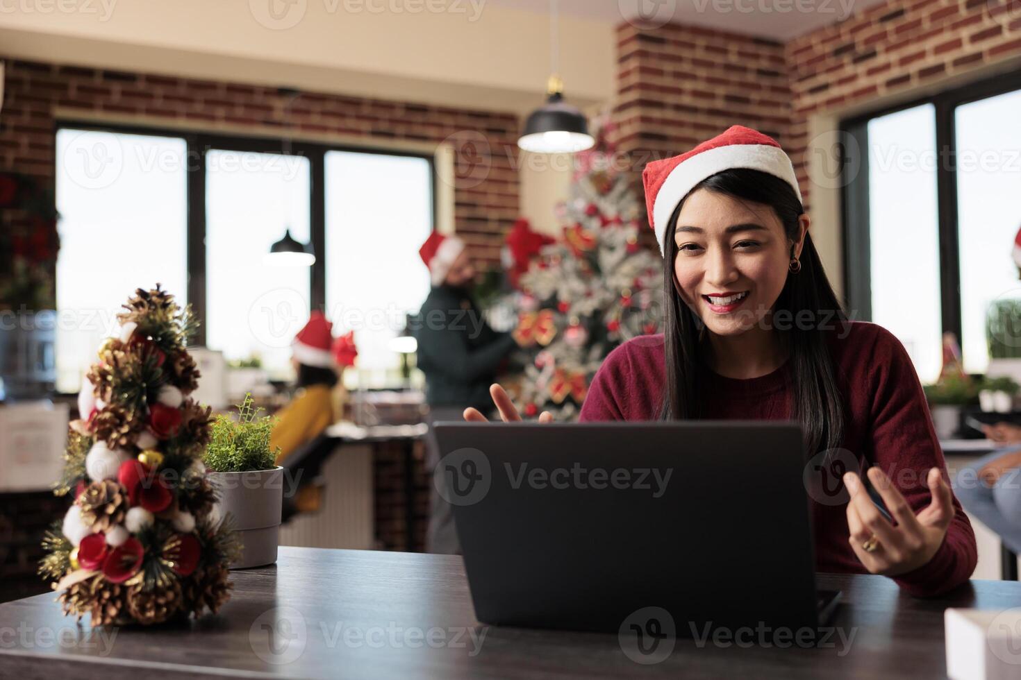 Smiling employee wearing santa hat managing project in teleconference using laptop in xmas decorated office. Cheerful woman worker having online communication at workplace during new year holiday photo