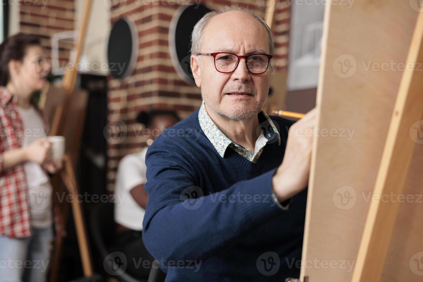 Pensive senior sitizen wearing glasses holding pencil drawing on canvas. Retired man attanding group art class in community centre to develop creative skills, spend time and socialize with others photo