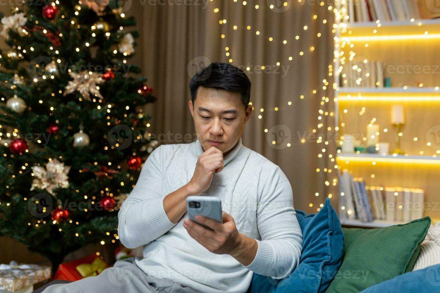 Serious thinking man reading bad message from smartphone sitting on sofa near Christmas tree in living room asian celebrating new year and holidays. photo