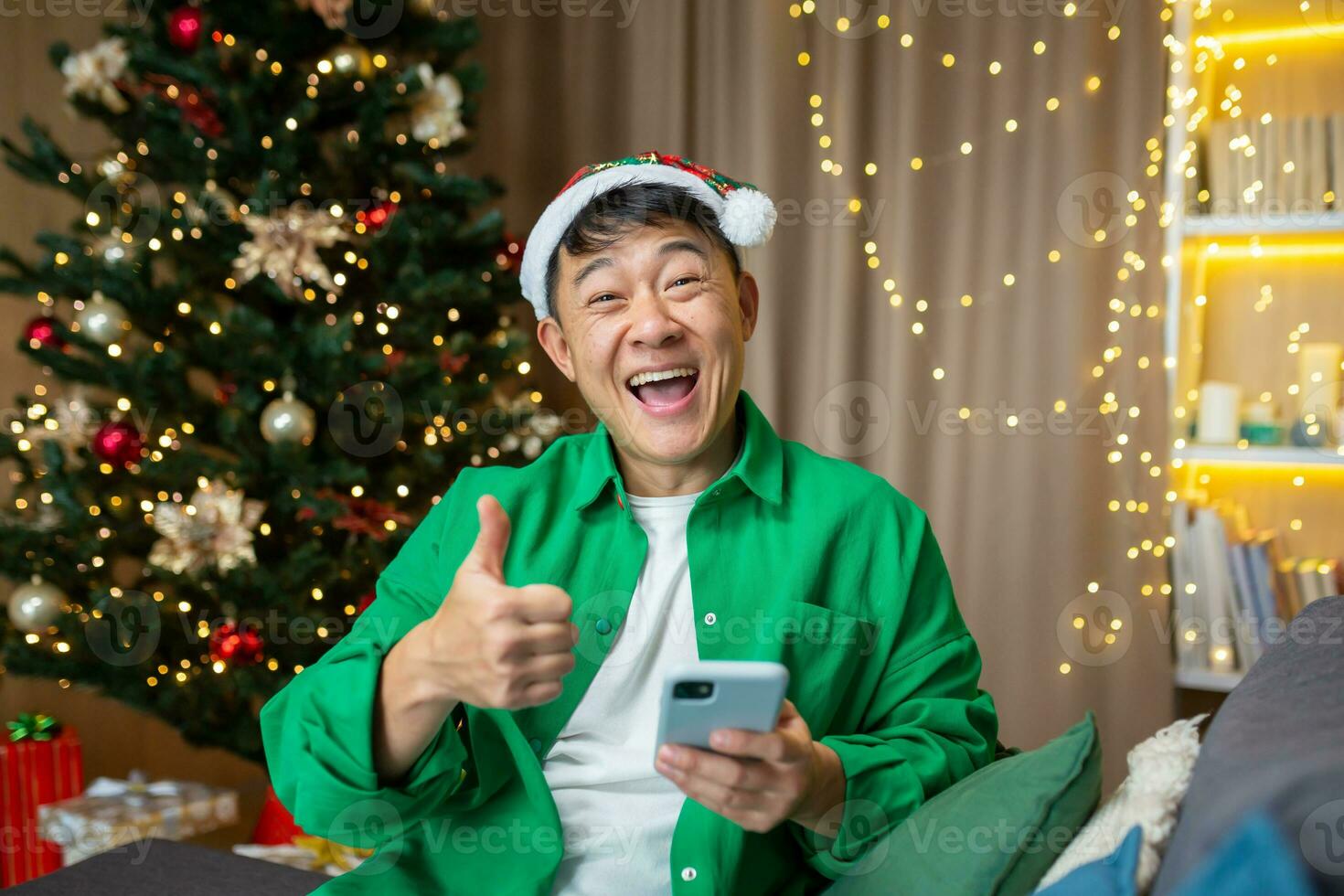 A young handsome Asian man holds a phone in his hands, is happy, received good news on the eve of Christmas and New Year holidays. Super points, Sitting on the sofa near the Christmas tree. photo