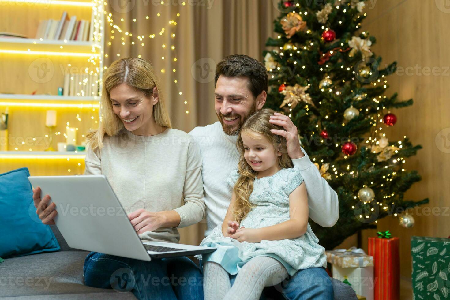 Happy family home for Christmas, husband wife and little daughter sitting on sofa in living room near Christmas tree, looking laptop and smiling celebrating New Year holidays, choosing gifts online. photo