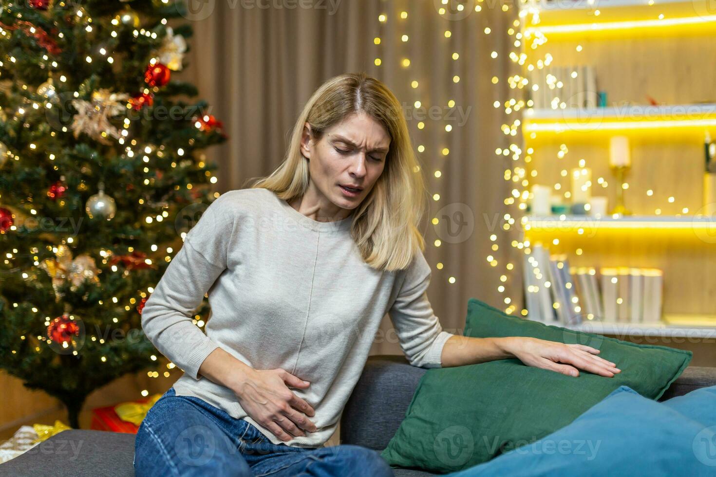 Woman abdominal pain and nausea on New Year's Eve sitting at home on the sofa against the background of a Christmas tree. Poisoning, heartburn, menstrual pain syndrome feels bad. photo