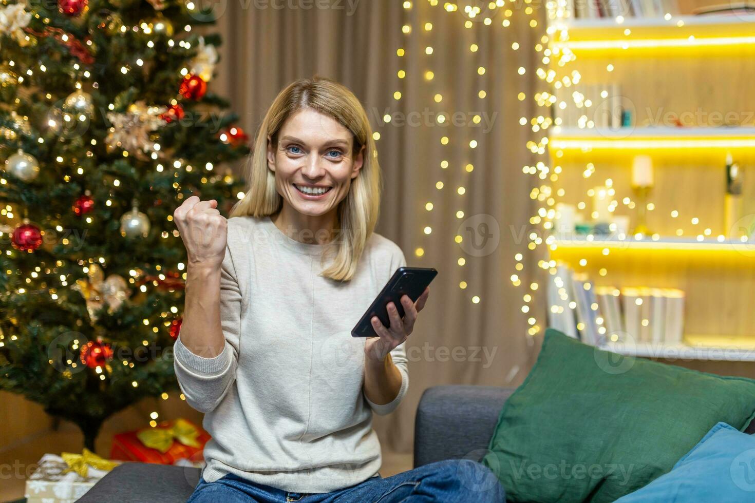 un contento joven mujer sostiene un teléfono en su manos, celebra un victoria, won un premio, recibido bueno noticias, hace un si mano. sentado a hogar en nuevo años víspera en el sofá cerca el Navidad árbol. foto