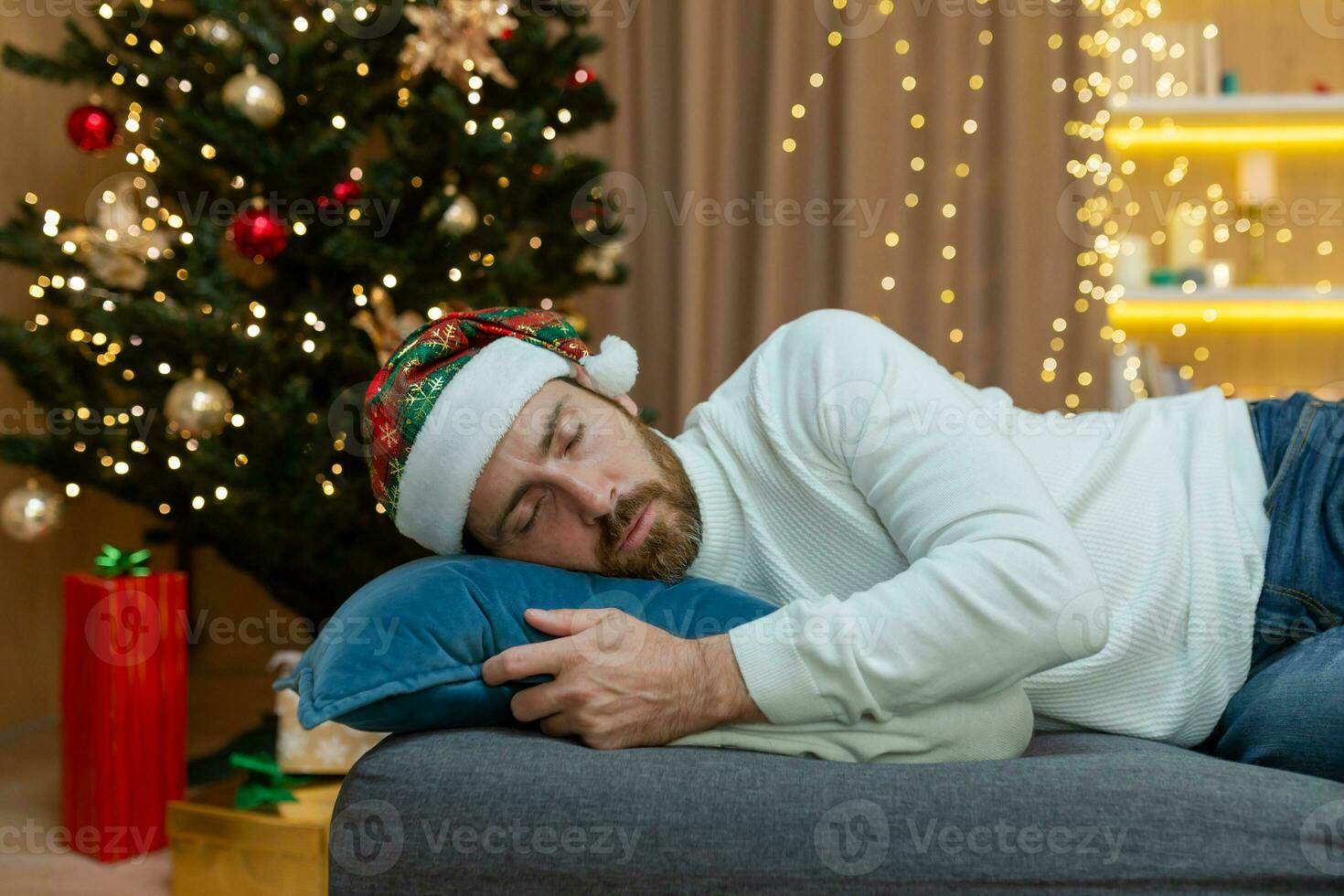 A man is tired for Christmas, wearing a New Year's hat, sleeping near the Christmas tree, sitting on the sofa at home in the living room after celebrating Christmas. photo