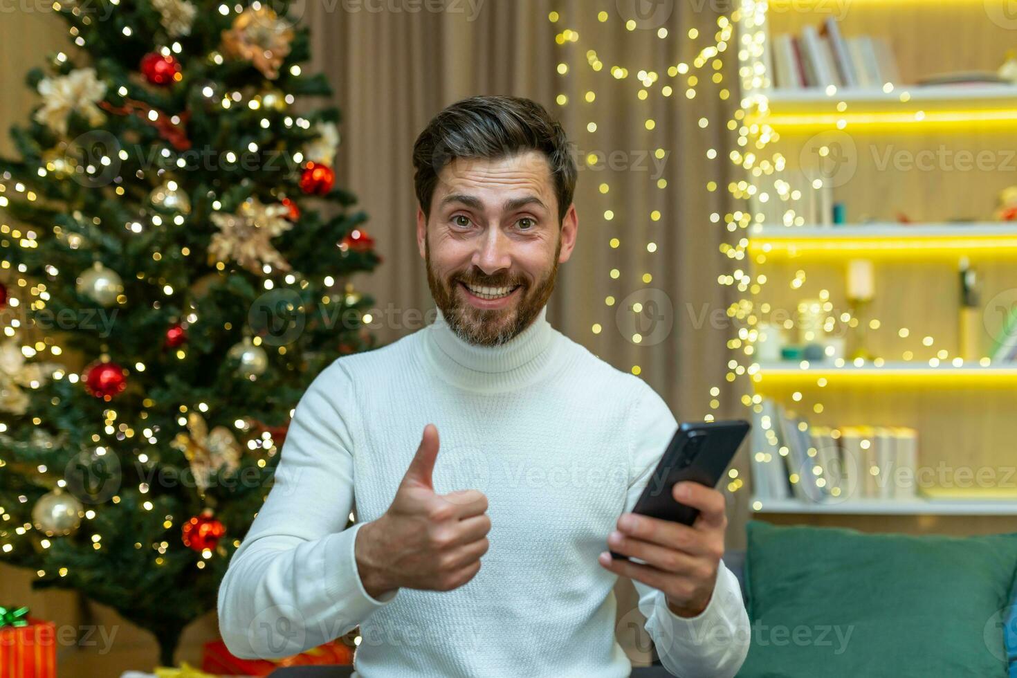 a man on Christmas looks at the camera and shows a finger up affirmatively holds a smartphone in his hands, sits at home on the sofa near the Christmas tree on New Year's holidays. photo