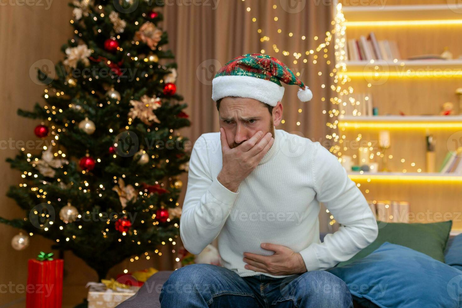Sick man at home for Christmas, sitting on the sofa in a New Year's hat has severe nausea and stomach pain. photo