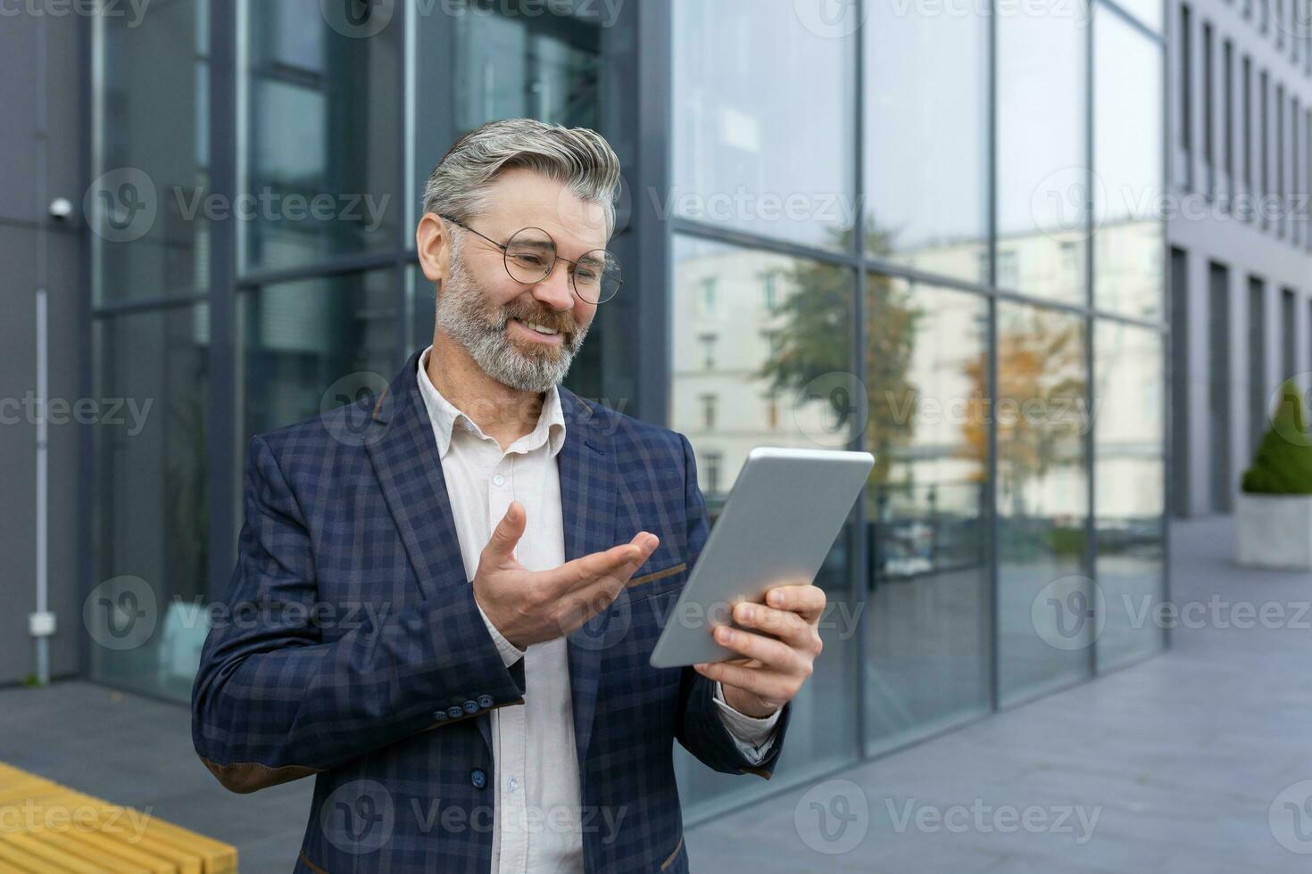 vídeo llamada remoto comunicación, maduro canoso empresario utilizando tableta computadora a hablar con amigos y colegas, jefe participación tableta fuera de oficina edificio al aire libre. foto