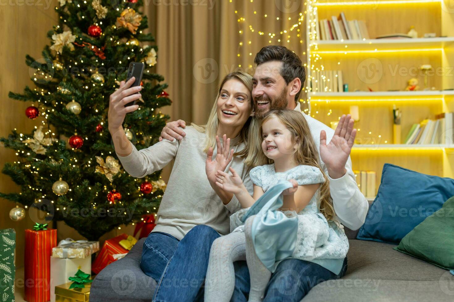 Happy family celebrating new year and christmas, husband wife and little daughter sitting on sofa in living room and talking with relatives on video call, using phone for remote communication. photo