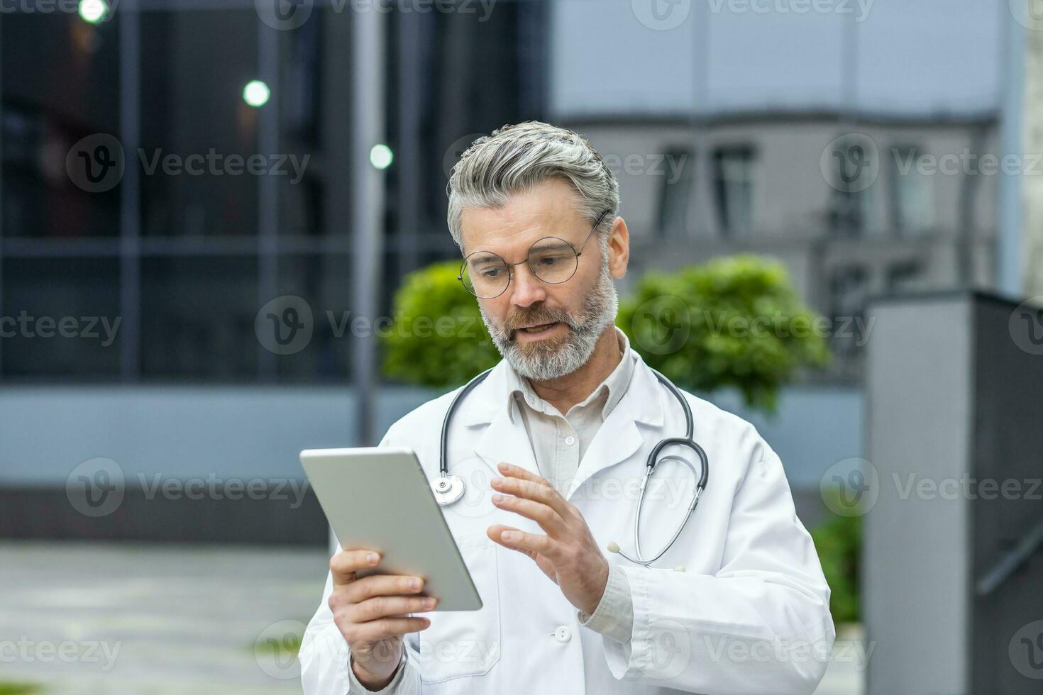 Serious mature doctor in medical gown outside modern clinic using tablet computer for video call, man with stethoscope consults patient online remotely. photo
