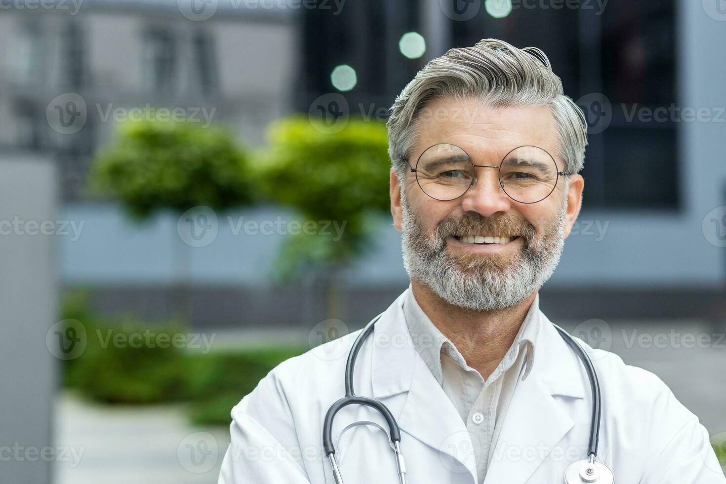 de cerca retrato de sonriente maduro médico en anteojos, mayor hombre sonriente y mirando a cámara fuera de moderno clínica al aire libre. foto