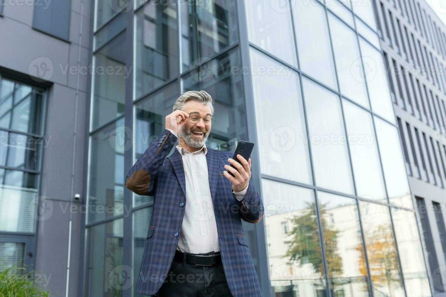 A happy senior grayhaired man in a suit and glasses stands outside an office, a skyscraper. Joyful reads a message on the phone, received good news, adjusts his glasses. photo