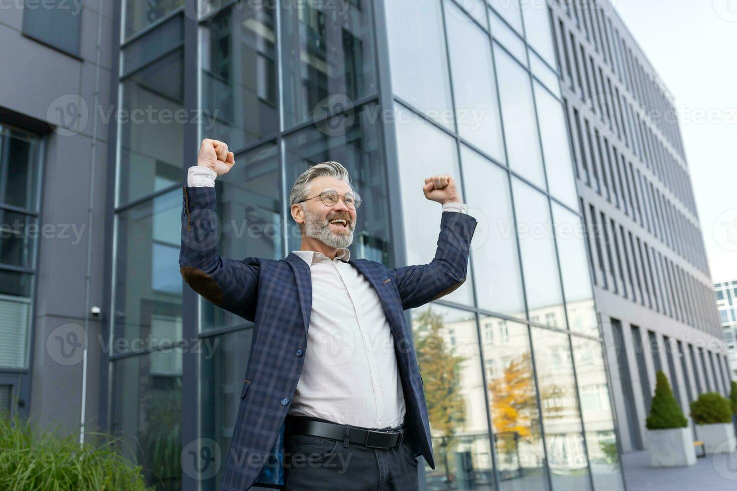 Successful and happy grayhaired boss outside office building rejoices success and victory, triumphs with hands up, businessman business owner in business suit and glasses, mature investor banker photo