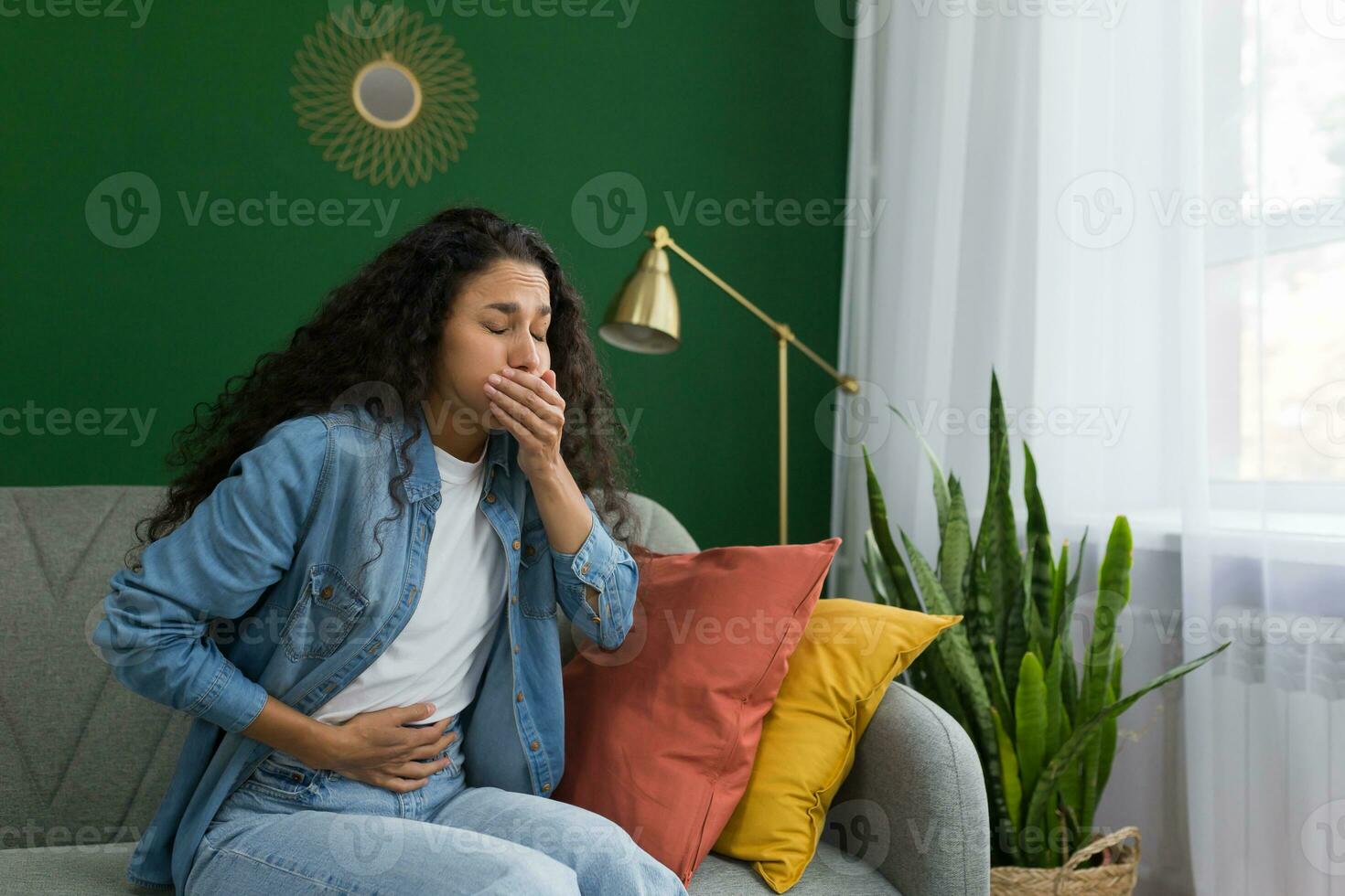 Young beautiful hispanic woman sick at home sitting on sofa with nausea and stomach ache sitting on sofa in living room near window with green wall photo