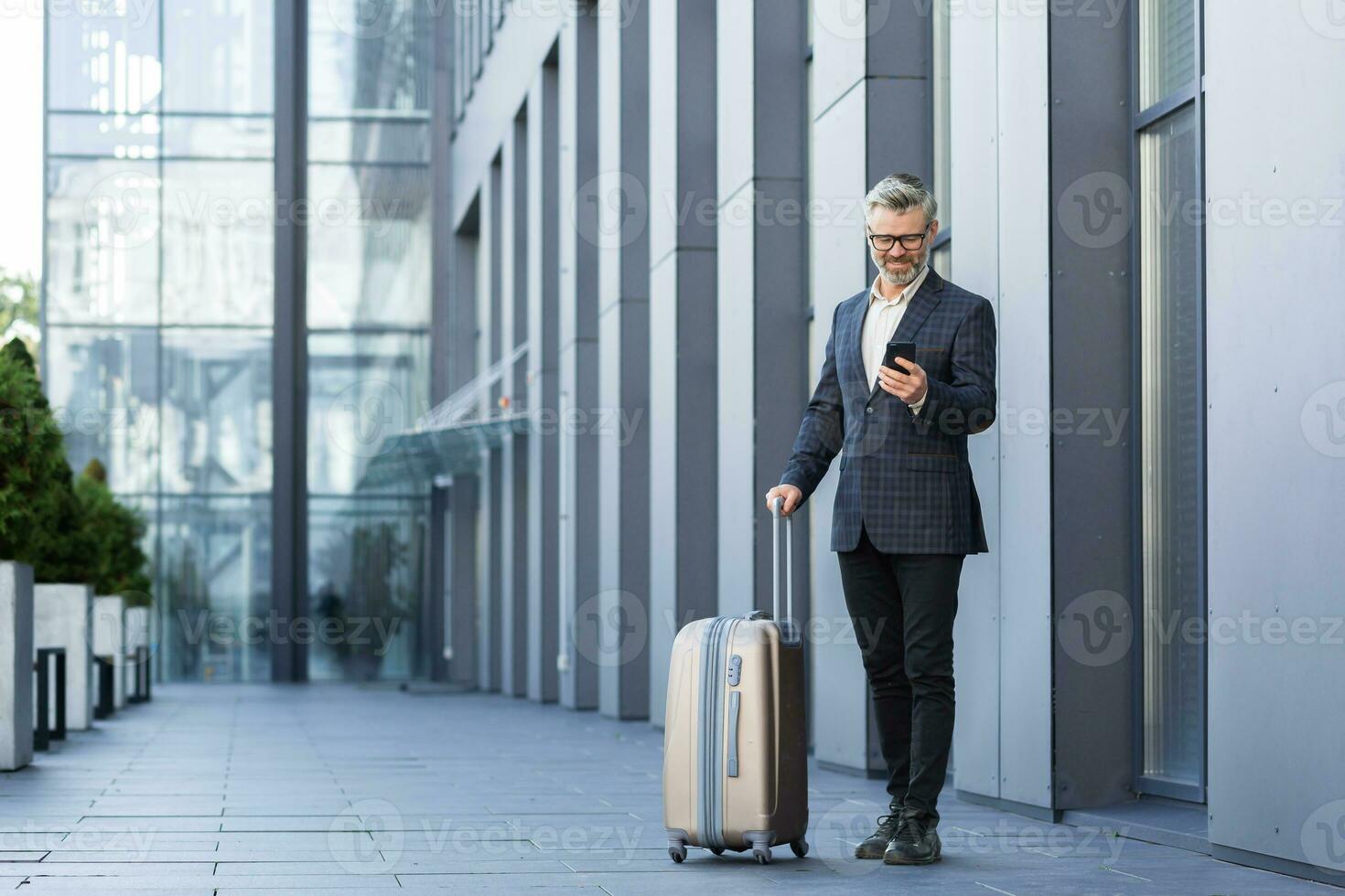 un hombre en un negocio traje con un grande viaje maleta y un teléfono es mecanografía un mensaje y reserva un Taxi, un jefe en un negocio viaje, un maduro empresario fuera de un grande hotel. foto