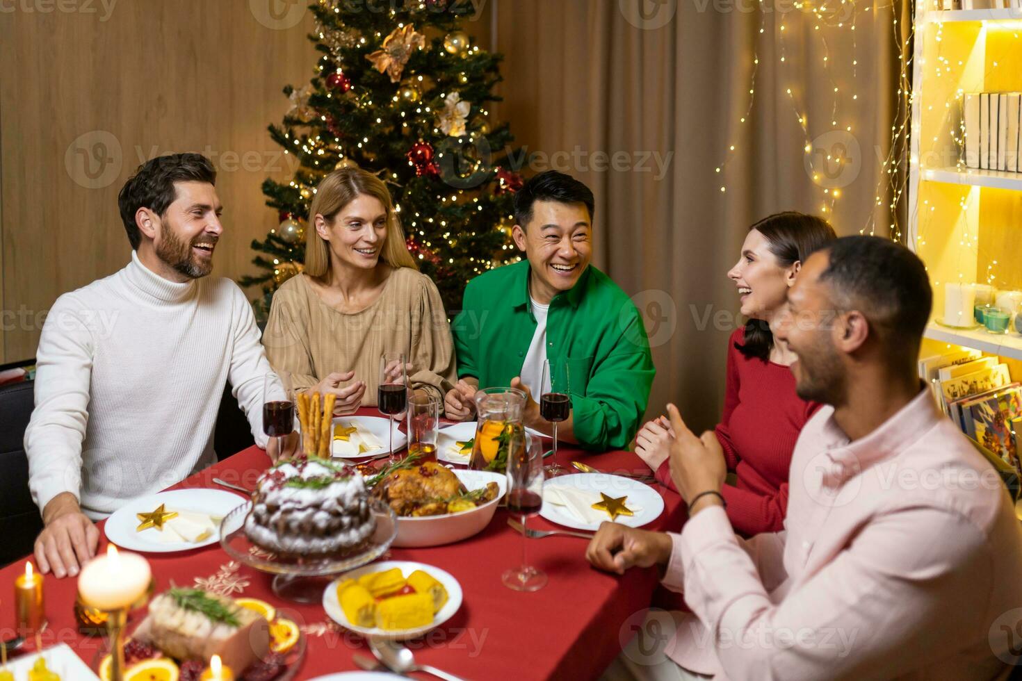 Happy young interracial people celebrating new year at home at festive table near Christmas tree. They hold glasses in their hands, laugh, communicate, rejoice photo