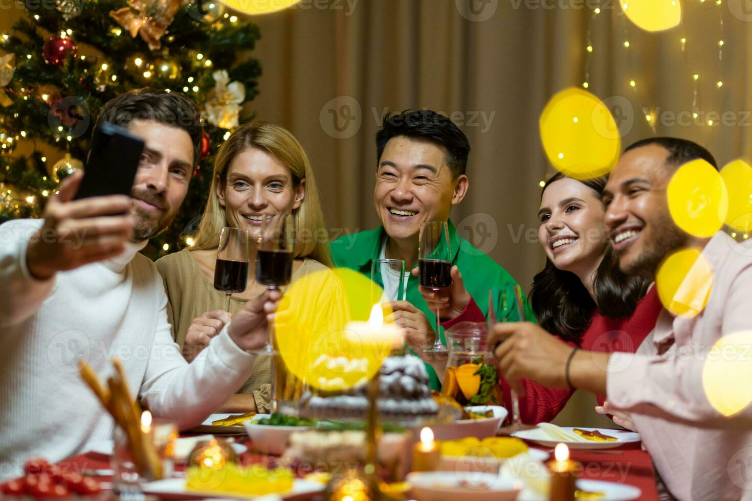 A company of interracial people celebrating the New Year together, sitting at the festive table. The man is holding the phone, everyone is talking on a video call with friends, greeting, smiling. photo